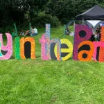 Photograph of large freestanding letters spelling "Play in the Parks" on a grass slope, each letter painted in bold, bright colours — pink, purple, turquoise, orange, red and yellow — with decorative dot and pattern designs on a black background. Behind the letters, families and children are gathered near a black gazebo, with dense green trees beyond. A cheerful, summery outdoor event scene.