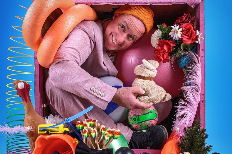 Promotional photograph of a grinning white man in a pink suit jacket and orange beanie hat, crammed into a pink wooden box overflowing with random objects. He is holding a small teddy bear. Surrounding him in the box: a pink exercise ball, a football, a bouquet of red and white roses, a foam pool noodle, a Slinky spring, a rubber chicken, a toy yellow school bus, a small plastic helicopter, a bundle of colourful pencils, pink feather boa, a small Christmas tree, and various other assorted tat. Blue background. He looks delighted with himself.