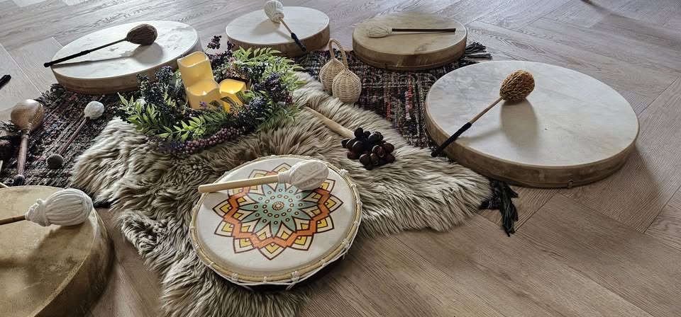 Photograph of a collection of frame drums and percussion instruments arranged on a sheepskin rug and patterned textile on a light wood herringbone floor. In the foreground, a drum decorated with a painted mandala in orange, gold, teal and green with a beater resting on it. Behind it, three larger plain hide frame drums with beaters. Between them, a small glowing LED candle, sprigs of dried foliage with dark berries, wicker rattles, and seed pod clusters. A gentle, ceremonial arrangement.