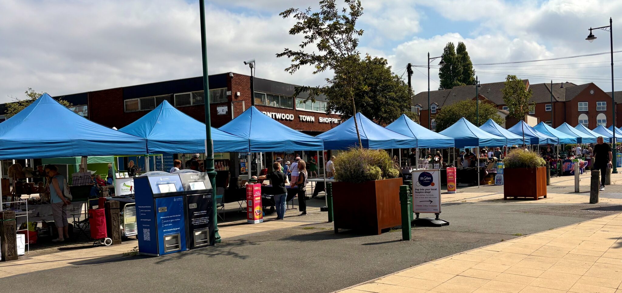Photograph of Burntwood Town Centre outdoor market on a sunny day. A long row of sky-blue gazebo stalls stretches across the town square in front of Burntwood Town Shopping Centre. Shoppers browse the stalls, with large rust-brown and dark metal square planters lining the walkway. A St Giles hospice charity sign is visible among the stalls. Trees and residential rooftops are visible in the background under a partly cloudy blue sky.